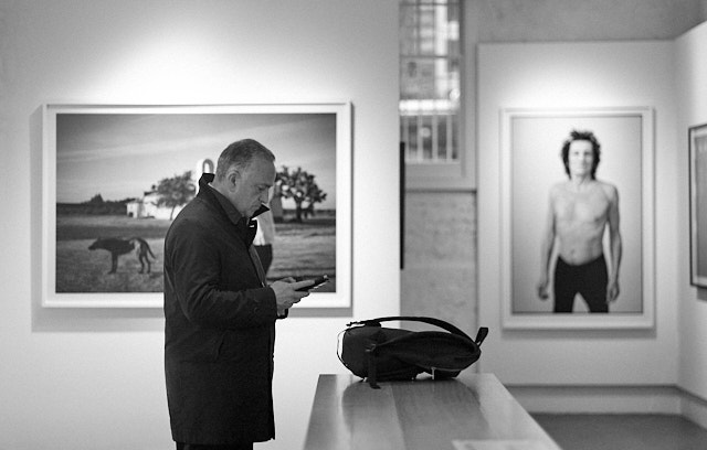 Charlie Gray exhibition in the Leica Gallery London. A gentleman checks his phone while Ronnie Wood poses in the background. Leica M10-R with Leica 90mm APO-Summicron-M ASPH f/2.0. © Thorsten Overgaard. 